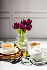 Green tea in a white porcelain cups and teapot, tulips in a glass vase, mimosa flowers, light background. Tea party.