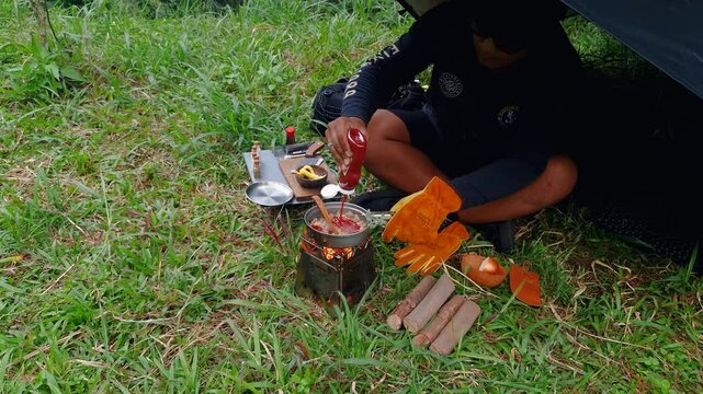 A male adventurer is cooking sauce to make a burger under a flysheet in the dense forest. The concept of bushcraft camping in a dense pine forest for adventure and cooking with simple equipment