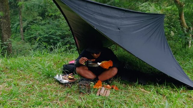 A male adventurer is cooking sauce to make a burger under a flysheet in the dense forest. The concept of bushcraft camping in a dense pine forest for adventure and cooking with simple equipment
