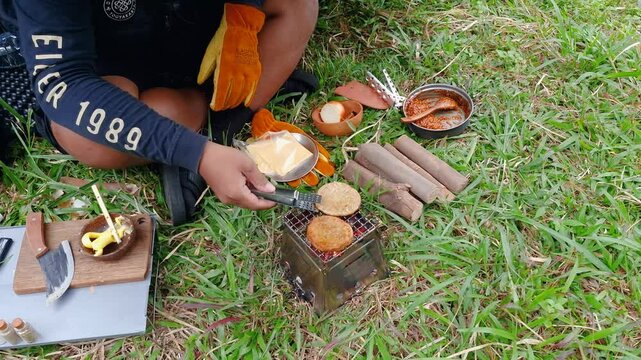 A male adventurer is cooking sauce to make a burger under a flysheet in the dense forest. The concept of bushcraft camping in a dense pine forest for adventure and cooking with simple equipment