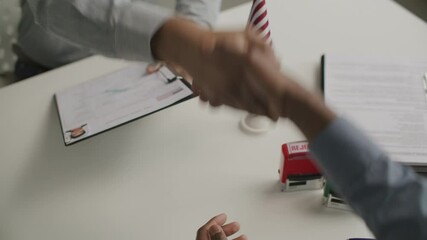 Hands of visa officer passing clipboard with approved application form and shaking hands with applicant at desk with American flag on it. Close-up view