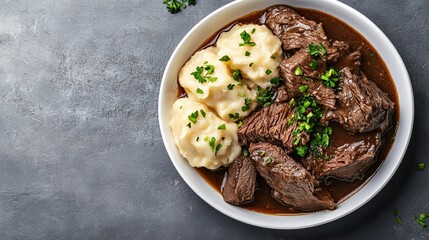 Sauerbraten meal served in a white bowl contains tender meat, rich gravy, and dumplings, garnished with fresh parsley on a textured gray surface.
