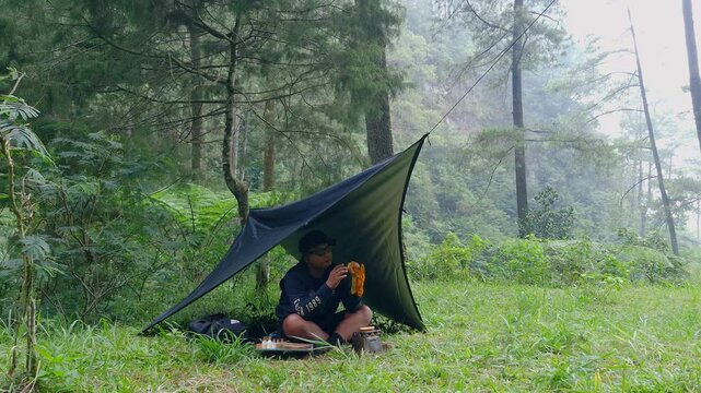 Male adventurer enjoying his homemade burger inside a flysheet in a pine forest area. Asian man's bushcraft camping concept in a quiet and beautiful pine forest