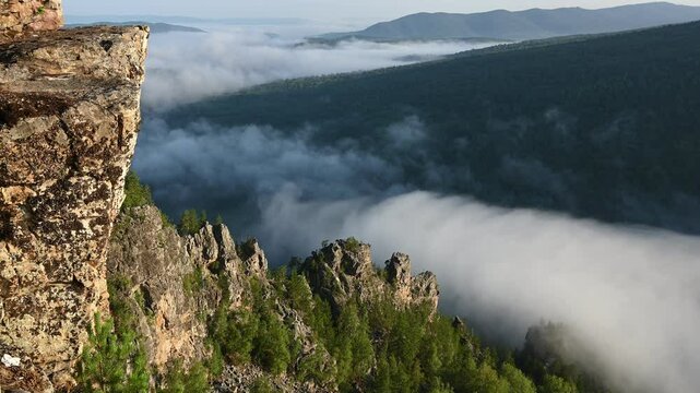 Ural mountains landscape. Aygir rocks. Clouds flow over a mountain valley, creating a stunning view from a cliff viewpoint surrounded by nature's beauty