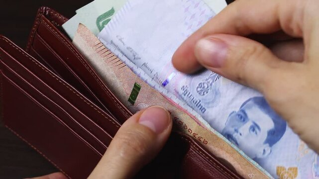 Close-up view of female hands holding wallet full of Thailand cash money, counting Thailand baht notes (THB) in the brown leather wallet. Wealth and success, financial profit and security.