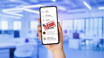 Hand holding smartphone displaying scam alert warning over blurred messaging app. Concept of cyber fraud, online scam detection, phishing attempts, digital security awareness, and identity theft preve