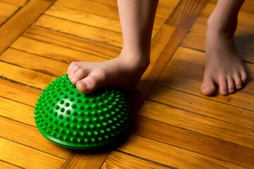 A child’s bare foot presses on a green spiky massage ball on a wooden floor, promoting relaxation or foot therapy. Perfect for wellness, health, or pediatric care themes.
