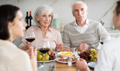 Mature spouses sitting at table, enjoying food and wine, chatting talking laughing with adult son and his girlfriend. In homely atmosphere male and female pensioner celebrate anniversary with guests