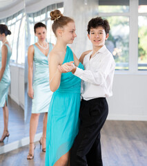 Obraz premium Couple of positive interested teenage dancers, boy and girl preparing for performance, rehearsing ballroom dance in spacious classroom while female teacher observing from background..