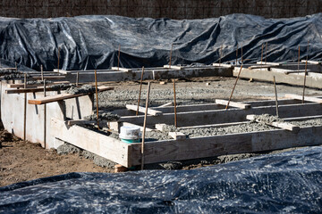 Construction site, new house at foundation stage, wood forms and rebar, ready for cement pour, black plastic tarps covering dirt piles, sunny winter day in housing development
