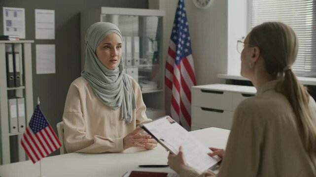 Young muslim woman in hijab handing application form to female visa officer and answering questions during immigration interview at US embassy