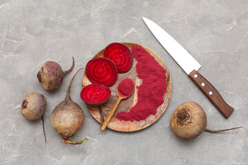 Wooden board with vibrant beetroot powder, spoon, knife, fresh beets and slices on grey background