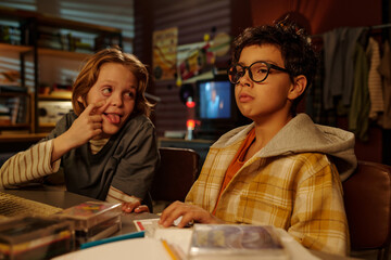 Two boys seated at desk in dimly lit bedroom, with one making goofy face while other looks serious. Room filled with books, posters, and personal items