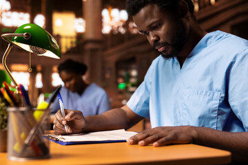 African american medical student creates a summary of class notes to learn easily for an exam, collecting information from the library to complete his knowledge. Future doctor at desk.