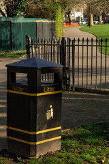 A black trash bin with yellow stripes in the park. In the background, there is an iron fence and a path.