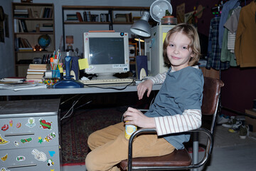 Portrait of young boy with long hair sitting at desk in a cozy, organized bedroom, holding a drink...
