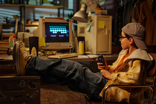 Young boy wearing glasses engrossed in playing video game on retro computer while sitting in cozy room filled with vintage decor and gaming equipment