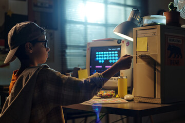 Young kid using an old computer in a dimly lit room, wearing a cap and large glasses, with a warm ambient background featuring retro decor and posters on walls