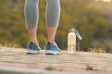Woman stands in athletic gear on a paved path near a water bottle during evening exercise in a natural setting