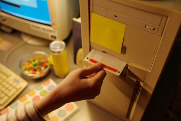 Hand inserting floppy disk into old desktop computer tower with yellow note and candies nearby in soft light. Retro setup featuring vintage technology and personal touch