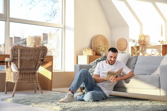 Handsome young man reading book on floor in stylish living room with big window