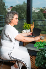 Beautiful woman works on laptop sitting in cozy room near window with cup. Blogger. Freelancer. Employment of retirees. Remote working.	