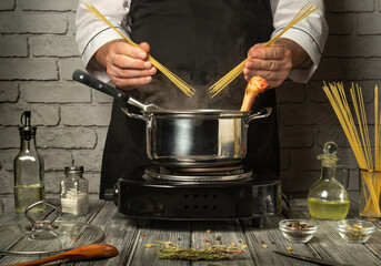 A chef in a black apron prepares pasta, holding dry spaghetti and a piece of chicken above a boiling pot. Fresh ingredients and spices decorate the kitchen countertop