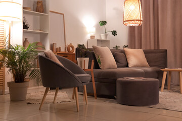 Interior of modern living room with grey sofa, armchair, pouf and glowing lamps at evening
