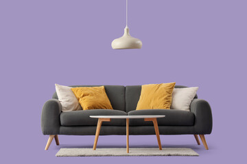 Cozy black sofa with cushions, coffee table and lamp on purple background