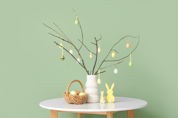 Vase with tree branches, Easter eggs, bunnies and wicker basket on table near green wall, closeup