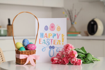 Basket of Easter eggs with greeting card and beautiful tulip flowers on table in festive kitchen