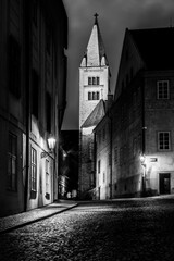 A nighttime view from Jirska Street reveals the Bell Tower of St. George's Basilica. Historic buildings frame the cobblestone street, illuminated by vintage street lamps.