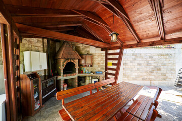 Wooden gazebo with tables and benches near the barbecue oven and kitchen cabinets on the wall