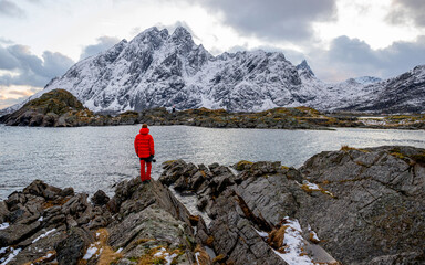 A hiker is standing on a rock in front of a lake with snow-covered mountains on the background, Lofoten, Norway