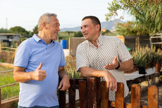 Neighbor conversation. Two smiling men breezily chatting near wooden plank fence of rural house on sunny summer day - Powered by Adobe