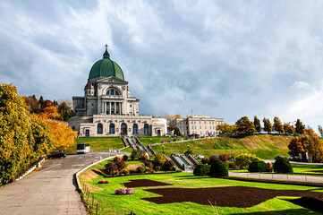 A stunning view of a grand basilica with its green dome, surrounded by autumn foliage. The foreground features a beautifully landscaped garden under a dramatic cloudy sky.