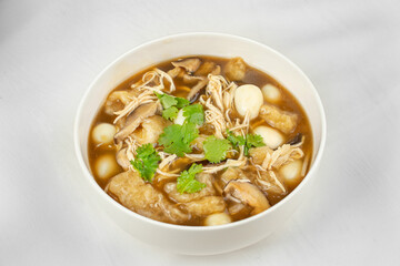 This image shows a white bowl filled with a dark broth soup containing shredded chicken, mushrooms, quail eggs, fried tofu, and garnished with fresh cilantro.