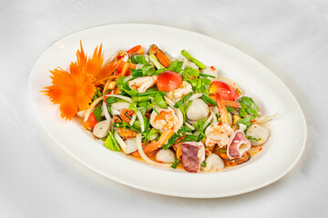 This image shows a white oval plate with a seafood salad featuring shrimp, squid, fish balls, onions, tomatoes, and herbs, garnished with a carved carrot flower.