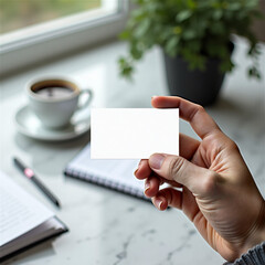 Obraz premium Hand holding blank business card mockup in cozy workspace with coffee cup, notebook, pen and houseplant. White card template against marble table background. Perfect for branding presentation or ident