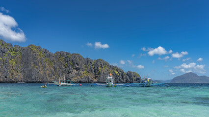 Fototapeta premium Traditional Filipino bangka boats are anchored in the aquamarine ocean. The canoe is floating. Green vegetation on the steep slopes of coastal cliffs. Blue sky, clouds. Philippines. Palawan.Bacuit Bay