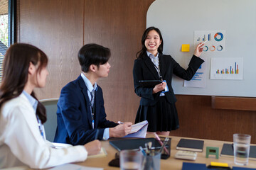 Confident young businesswoman presenting marketing analytics on a whiteboard while startup team discusses strategy, collaboration, and financial growth in a modern office