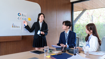 Confident young businesswoman presenting marketing analytics on a whiteboard while startup team discusses strategy, collaboration, and financial growth in a modern office