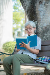 Fototapeta premium Elderly asian woman wearing glasses and headphones around her neck is sitting on a bench in a park, enjoying reading a book in the shade of a tree, with a colorful scarf resting beside her