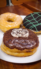 Three delicious-looking donuts on a white plate on brown table. The donut in the foreground is the focal point, with a dark chocolate coating topped with cream and sprinkled with chocolate shavings