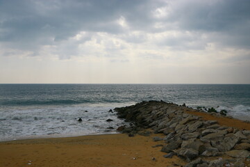 This image captures a coastal scene with a sandy beach, ocean waves, and a cloudy sky. The shoreline has large rocks acting as a barrier