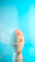 Single white egg sits in a white egg cup against a vibrant light blue backdrop, creating a simple yet visually appealing composition with a focus on breakfast and healthy eating, easter