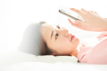 A young and beautiful woman, dressed in pink, is lying on a bed in a sunlit room, smiling happily while looking at her smartphone