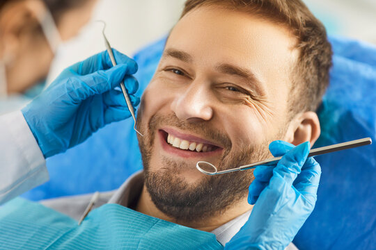Happy man undergoing dental check up during visit of dentist, smiles toothily, showing white perfect tist. Unrecognizable dentist using dentistry instrument to examine patients oral cavity