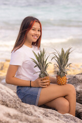 Beautiful teenager girl sitting on the stone, holding two whole pineapples. Smiling face. Organic fruit concept. Bali.