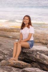 Happy slim teenager girl sitting on the stone on the beach. Summer vacation in Asia. Enjoy traveling to Bali island.